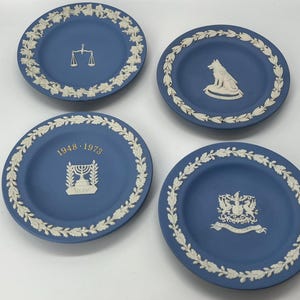 May include: Four blue decorative plates with white relief designs. Each plate features a different emblem: scales, a wolf, a menorah with the dates 1948-1973, and a coat of arms. The plates have a white leaf border.