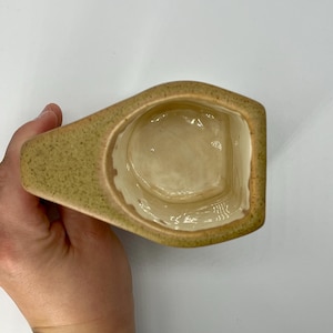 Peut inclure: Une tasse en céramique de forme unique, avec un extérieur vert clair et un intérieur crème. La tasse a une anse et une large ouverture, parfaite pour contenir des boissons. La tasse est tenue par une main.