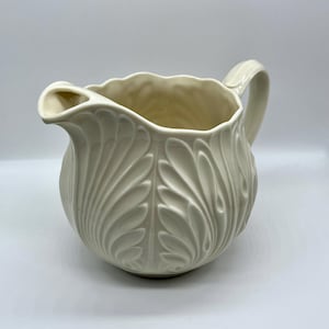 Puede incluir: Jarra de cerámica color crema con diseño de hojas. La jarra tiene un cuerpo redondeado, un asa curva y un pico para verter. La superficie es lisa con un patrón de hojas en relieve. El borde de la jarra tiene un borde festoneado.