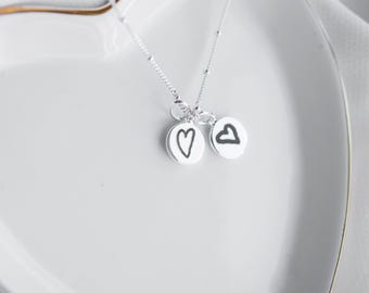 Könnte beinhalten: Eine zarte Silberkette mit zwei kleinen, runden Anhängern. Ein Anhänger hat ein handgezeichnetes Herz, der andere den handgezeichneten Buchstaben "b". Die Halskette wird auf einem weißen, herzförmigen Teller präsentiert.