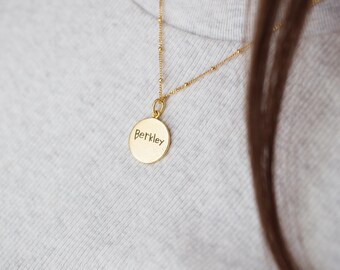 May include: A gold-colored necklace with a round pendant. The pendant has the name "Berkley" engraved in a handwritten style. The necklace is worn over a gray sweater.