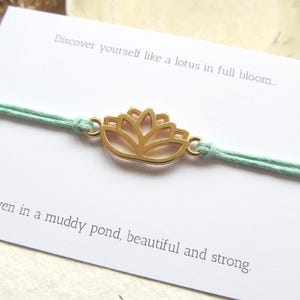 Puede incluir: Una pulsera con un dije de flor de loto dorada y un cordón verde claro. La pulsera se muestra en una tarjeta blanca con el texto "Discover yourself like a lotus in full bloom...even in a muddy pond, beautiful and strong."