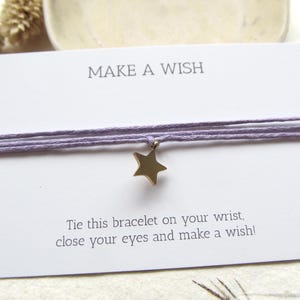 Puede incluir: Una pulsera de hilo lila con un colgante de estrella dorada, presentada en una tarjeta blanca. La tarjeta dice "MAKE A WISH" e incluye instrucciones para atar la pulsera y pedir un deseo.