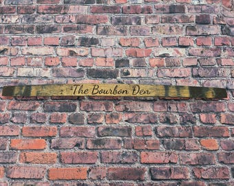 May include: A wooden sign with the text "The Bourbon Den" engraved on it. The sign is made from a piece of wood with dark stripes and is set against a brick wall.