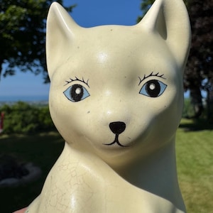 May include: A cream-coloured ceramic cat figurine with blue eyes and black details. The cat has a slightly cracked, aged appearance. The figurine is outdoors with a blue sky and green trees in the background.