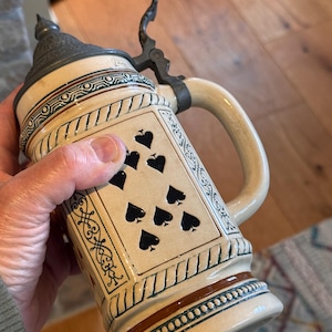 Puede incluir: Un jarro de cerveza de cerámica vintage con tapa y asa metálicas con bisagras. El jarro presenta una base de color crema con un diseño de picas negras. Los adornos decorativos en marrón y verde acentúan el diseño. El jarro se sostiene.