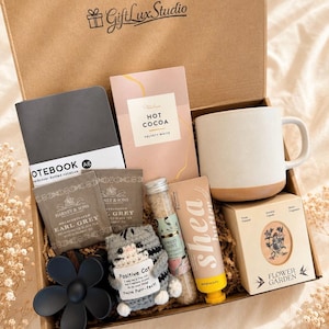May include: A gift box filled with a variety of items, including a gray notebook, hot cocoa, a mug, tea bags, a small cat plushie, hand cream, a candle, and a black flower-shaped item. The box is brown with the text "Gift Lux Studio".