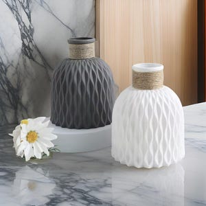 Peut inclure: Deux vases décoratifs. L'un est gris, l'autre blanc. Les deux ont un motif texturé en forme de vagues sur le corps et un col enroulé de corde. Une fleur blanche avec un centre jaune est à proximité.