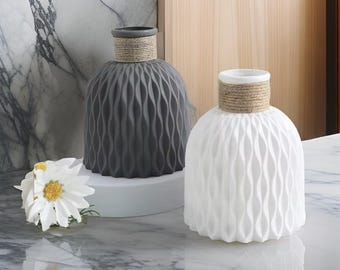 May include: Two decorative vases. One is gray, the other is white. Both have a textured, wave-like pattern on the body and a rope-wrapped neck. A white flower with a yellow center sits nearby.