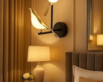May include: A gold bird-shaped wall sconce with two lights, mounted on a black wall in a bedroom setting. The sconce is next to a bedside table with a lamp and a vase of flowers.
