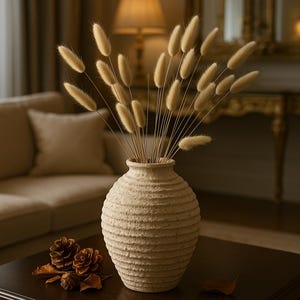 Peut inclure: Un vase beige texturé rempli d'herbe de queue de lapin séchée. Le vase est posé sur une table marron foncé, avec des pommes de pin et une feuille. L'arrière-plan comprend un canapé, une lampe et un miroir.