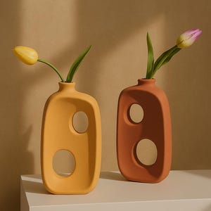 以下が含まれることがあります： 切り抜きデザインのセラミック花瓶2つ。左の花瓶は黄色で、1本の黄色のチューリップが入っています。右の花瓶はオレンジ色で、1本のピンクのチューリップが入っています。
