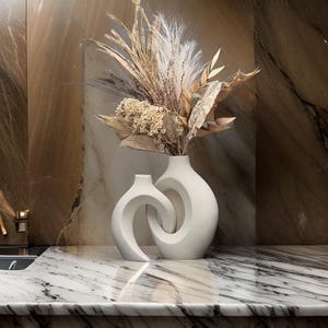 May include: Two white ceramic vases with an abstract design. The vase on the right is round and the vase on the left is shaped like a wave. The vases are filled with dried flowers.
