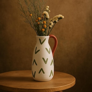 Peut inclure: Un vase en céramique blanc avec une poignée rouge et un motif de chevrons verts, rempli de fleurs et de feuillage séchés. Le vase est posé sur une table ronde en bois sur fond marron. Un élément de décoration intérieure.