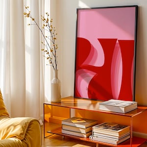 Könnte beinhalten: Ein gerahmter abstrakter Kunstdruck mit schwarzem Rand, der rote und rosa Formen zeigt. Das Kunstwerk wird auf einem orangefarbenen Konsolentisch mit Büchern und einer weißen Vase mit einem Zweig mit gelben Knospen ausgestellt. Ein gelber Sessel befindet sich im Vordergrund.