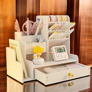Puede incluir: Un organizador de escritorio de madera blanca con múltiples compartimentos para artículos de papelería. Cuenta con un cajón, estantes y ranuras para bolígrafos, marcadores y otros suministros. También se ve un pequeño temporizador digital y objetos decorativos.