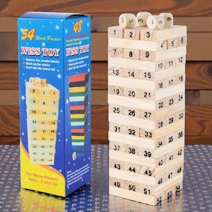 Puede incluir: Un juego de apilamiento de bloques de madera con 54 piezas, numeradas del 1 al 54, y cuatro dados. La caja dice "Wiss Toy" y "Equilibra los bloques de madera, Apila las historias, No dejes que la torre caiga". El juego es para mayores de 5 años.