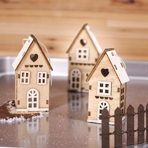 Op de afbeelding: Drie kleine houten huisornamenten met hartvormige uitsparingen en witte accenten. Elk huis heeft een driehoekig dak en een kleine deur. Een miniatuur bruin hek staat op de voorgrond.