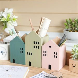 May include: Three pastel-coloured wooden house-shaped desk organisers. The organisers are painted in teal, light green, and pink, each with black window and door cutouts. Pens, pencils, and papers are inside. A notebook, glasses, and a clock are also visible.