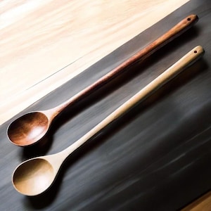 May include: Two wooden spoons, one dark brown and one light beige, are displayed diagonally. The spoons have long handles and rounded bowls. The dark brown spoon is positioned above the light beige spoon. Both spoons have a small hole at the end of the handle.