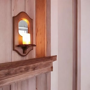 Op de afbeelding: Een houten wandkandelaar met een kaars en een kleine spiegel. De kandelaar is donkerbruin van kleur en is bevestigd aan een lichtbruine houten muur. De kaars is aangestoken en geeft een warme gloed.