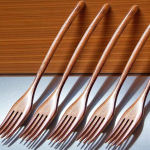 May include: Six wooden forks with a natural finish. The forks have a classic design with four tines and long, curved handles. The forks are arranged diagonally against a wooden background.