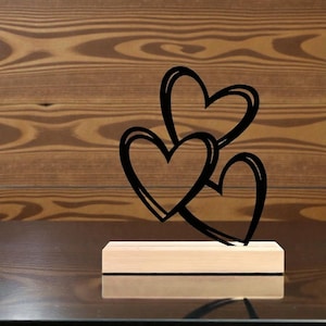 Puede incluir: Una escultura de metal negro con tres corazones entrelazados, montada sobre una base rectangular de madera. Los corazones tienen un estilo dibujado a mano, con un doble contorno. La base de madera es de color claro.