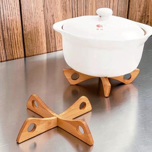 May include: A white ceramic pot with a lid featuring a small red logo and text, resting on a light brown bamboo cross-shaped trivet. A second identical bamboo trivet is positioned in the foreground, showcasing its four-legged design with circular cutouts.