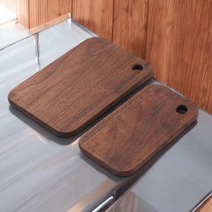 Puede incluir: Dos tablas de cortar rectangulares de madera con esquinas redondeadas y un agujero para colgar. Las tablas son de color marrón oscuro y parecen ser de madera dura. Se muestran sobre una superficie metálica brillante.
