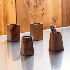 Peut inclure: Quatre porte-stylos en bois de différentes formes. Trois sont rectangulaires et un est trapézoïdal, contenant des stylos. Le grain du bois est visible, et les supports sont sur une surface réfléchissante. L'arrière-plan est un panneau de bois.