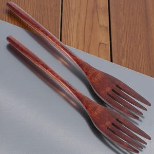 May include: Two wooden forks with a rich, reddish-brown finish. The forks are positioned diagonally on a gray surface, with a wooden background visible. The tines are slender and evenly spaced, suggesting a utensil for serving or eating.