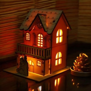 Puede incluir: Una maqueta de casa de madera iluminada, con un exterior naranja claro y un tejado de color claro. La casa tiene varias ventanas que brillan con luz cálida y un pequeño balcón. Una piña decorativa se encuentra a la derecha de la casa.