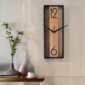 May include: A rectangular wall clock with a black frame and a wooden face. The clock face features a light brown vertical stripe flanked by darker brown wood. Black hands point to the numbers 12 and 6.