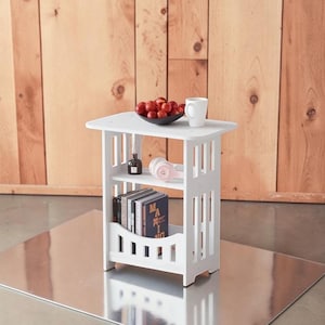 May include: A white, three-tiered side table with a square top and slatted sides. The top shelf holds a black bowl of red fruit and a white mug. The middle shelf has pink headphones, and the bottom shelf has books.
