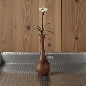 May include: A tall, wooden vase with a slender neck and bulbous base holds a single artificial flower. The flower has a white and yellow center with green leaves. The vase is a rich brown color, and the background is a wooden wall.