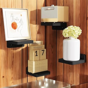 Puede incluir: Una exhibición de estantes flotantes negros montados en una pared de madera, cada uno con diferentes elementos decorativos. Los elementos incluyen una impresión artística enmarcada, un electrodoméstico beige y dorado, un jarrón blanco con flores y un calendario perpetuo de madera.