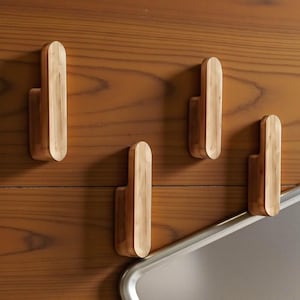Op de afbeelding: Vier houten wandhaken met een lichtbruine afwerking zijn gemonteerd op een houten oppervlak. De haken hebben een afgeronde bovenkant en een rechthoekige basis. Een zilverkleurig metalen frame is zichtbaar in de rechter benedenhoek.
