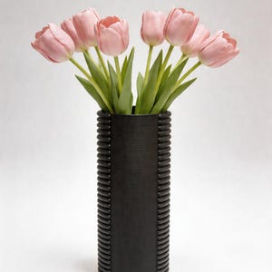 Könnte beinhalten: Eine schwarze, strukturierte Vase präsentiert einen Strauß rosa Tulpen mit grünen Stielen und Blättern. Die Vase, etwa 30 cm hoch, hat eine zylindrische Form mit vertikalen, gerippten Details. Die Tulpen blühen voll, ihre zarte rosa Farbe kontrastiert mit der dunklen Vase.