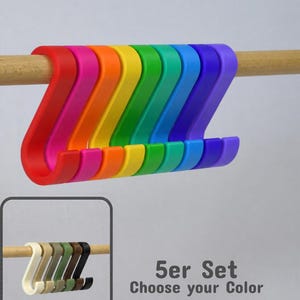 Könnte beinhalten: Ein Set aus S-förmigen Haken in Regenbogenfarben wie Rot, Pink, Orange, Gelb, Grün, Türkis, Blau und Lila an einer Holzstange. Ein kleiner Kasten zeigt ein Set in neutralen Farben mit dem Text 5er Set Choose your Color.