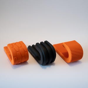 Könnte beinhalten: Drei Türstopper aus Gummi in Orange und Schwarz. Die orangenen Stopper haben strukturierte Oberflächen, einer mit einem ovalen Muster und der andere mit einem gerippten Design. Der schwarze Stopper hat ein gebogenes, geriffeltes Design.
