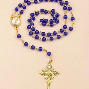 Puede incluir: Un rosario de vidrio azul con detalles dorados. El rosario presenta una cruz dorada con una representación de Jesucristo y un colgante redondo con una imagen de un ángel.