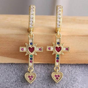 Puede incluir: Aretes de aro dorados con un dije de cruz colgante. La cruz está decorada con piedras preciosas de colores y un corazón rojo en el centro. Los aretes también tienen un pequeño dije de corazón que cuelga de la parte inferior.