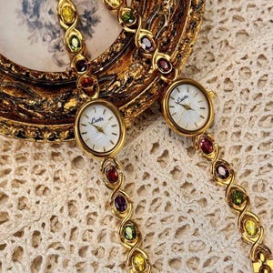 Puede incluir: Dos relojes de tono dorado con correas enjoyadas. Los relojes tienen esferas blancas con marcadores de hora en números romanos negros y la palabra "L'Autre" impresa en la esfera. Las correas son de metal dorado y tienen piedras de colores alternas.