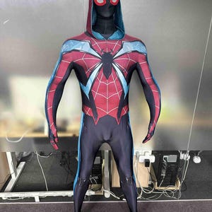 Può includere: Un costume da supereroe rosso, nero e blu, con un'emblema di ragno sul petto. Il costume include un cappuccio con una maschera ed è progettato per coprire l'intero corpo. La tuta ha un motivo a ragnatela ed è realizzata in materiale elastico.