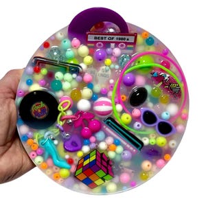 Puede incluir: Una bandeja redonda de resina transparente llena de cuentas de colores y abalorios con temática de los años 80. Incluye un Cubo de Rubik, gafas de sol, una cinta de cassette que dice "Best of 1980s" y un disco que dice "Rock & Roll".