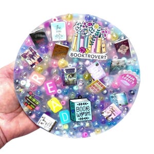 May include: A circular resin coaster with miniature book charms, colourful beads, and the word "READ" spelled out in individual letter charms. The coaster also features the text "BOOKTROVERT" and "BOOK WORM".