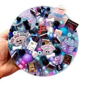 Puede incluir: Una pieza de arte redonda de resina con una variedad de dijes de libros en miniatura, cuentas y elementos decorativos. El diseño incluye el texto "LET ME READ", "BOOKISH GIRLIE" y "SPOOKY BOOK CLUB". La paleta de colores presenta azules, morados, rosas y negro.