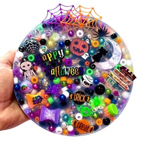 May include: A round Halloween-themed resin art piece with a variety of colorful beads, charms, and embellishments. The design includes a jack-o'-lantern, spiderwebs, a black cat, and the words "Happy Halloween", "Trick", and "Treat".
