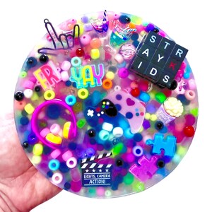 Puede incluir: Una colorida pieza de arte de resina redonda con cuentas, dijes y texto incrustados. El diseño incluye las palabras "FR" y "YAY", un mando de juegos, una claqueta de cine con "LIGHTS. CAMERA. ACTION!" y un bloque "STRAY KIDS".