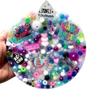 Op de afbeelding: Een rond kunstwerk van hars met diverse versieringen. Het bevat teksten als "Sprinkle Kindness" en "Be the Reason Someone Smiles Today!" in een mix van kleuren, waaronder roze, blauw en groen. Het stuk is gevuld met kralen en bedels.